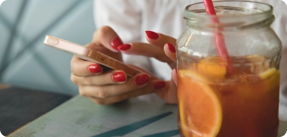 Eine Frau mit roten Nägelt hält ein Smartphone und tippt. Neben ihr steht ein Glas mit Orangensaft.